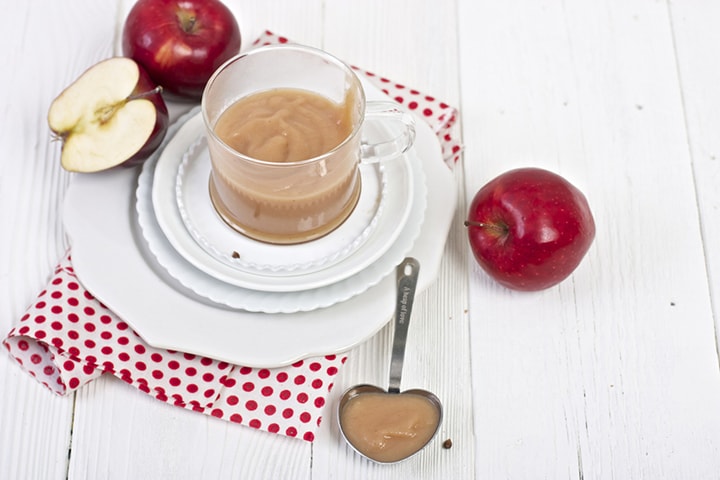 Image of a clear bowl filled with baby food puree with produce surrounding it and a spoon in front with a bite for baby.