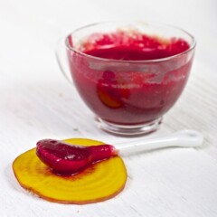 clear jar filled with red beet puree with a spoon sitting in front of it resting on a piece of golden beet.