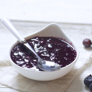 small white bowl filled with a blueberry baby food puree inside. bowl has a white and silver spoon resting on top and is sitting on a white surface