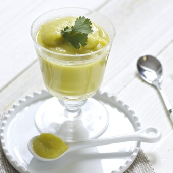 A clear jar filled wit a creamy yellow squash baby food puree. The jar is sitting on a white plate on a white wooden surface.