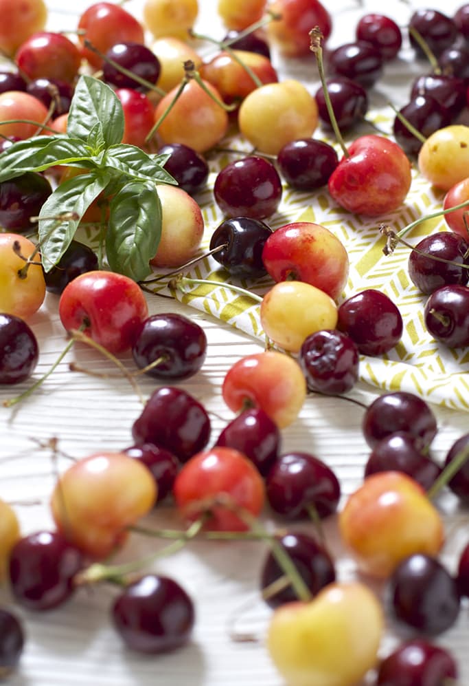 A bunch of sweet and tart cherries scattered across a white wooden board with a basil leaf in the middle.