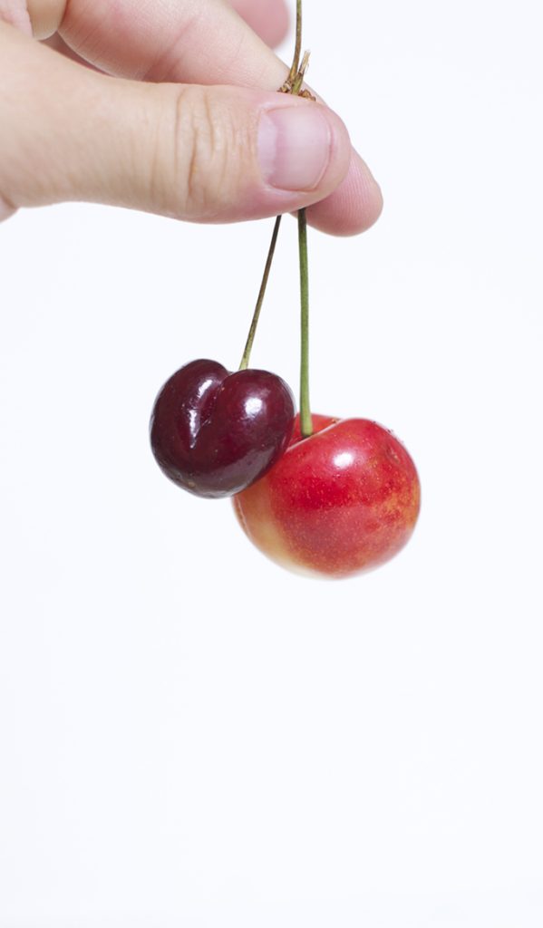 Side shot of fingers holding on to 2 cherries by the stems.