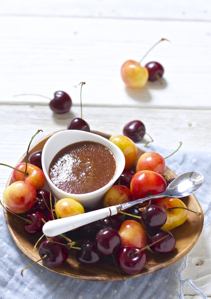 small white bowl sitting on a wooden plate that is filled with whole cherries. In the bowl is a thick red cherry baby food puree.