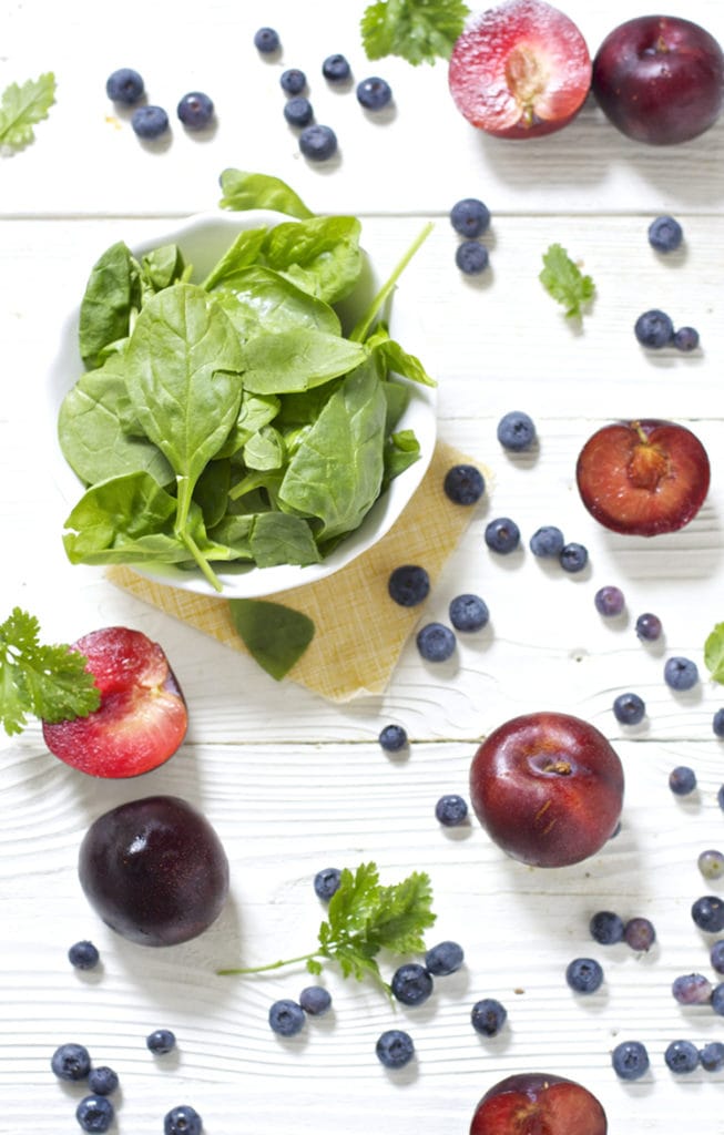 Scattering of produce - plumes, blueberries, spinach and cilantro all over a white board.