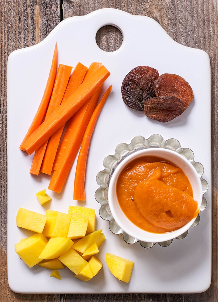 Image is of a white cutting board with chopped produce on it and one small bowl filled with a creamy and thick baby food puree.