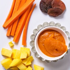 Image is of a white cutting board with chopped produce on it and one small bowl filled with a creamy and thick baby food puree.