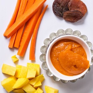 Image is of a white cutting board with chopped produce on it and one small bowl filled with a creamy and thick baby food puree.
