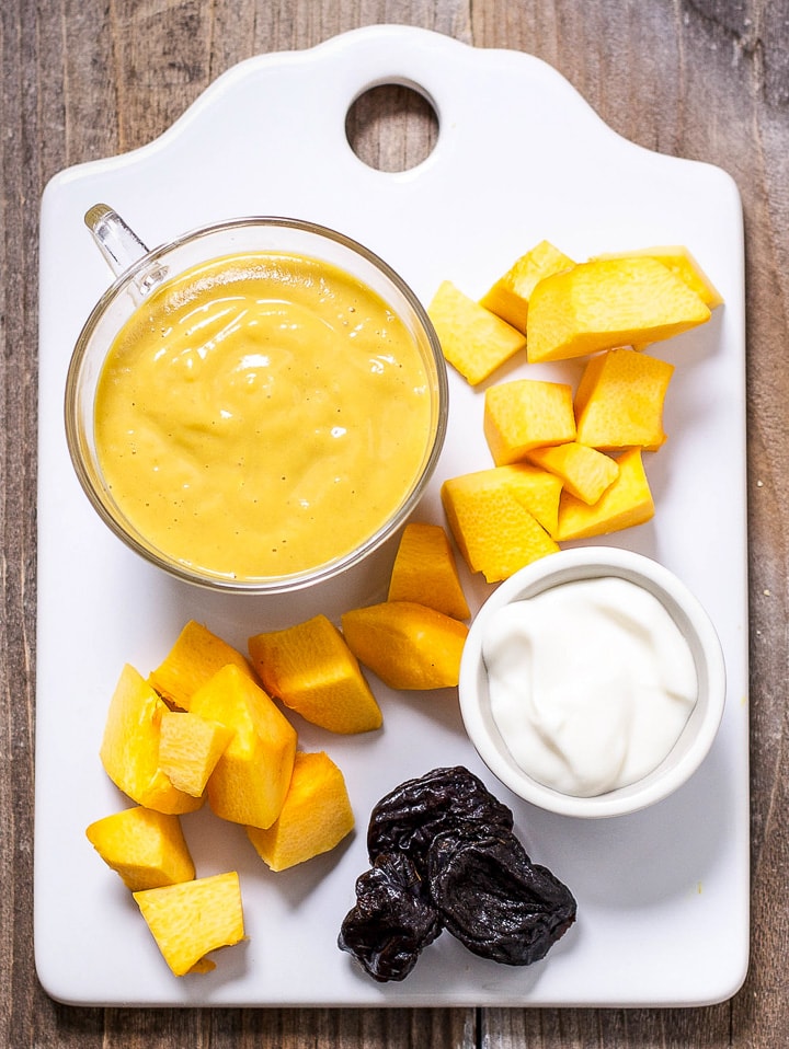 Image is of a white cutting board with produce scattered with a small bowl filled with baby food puree.
