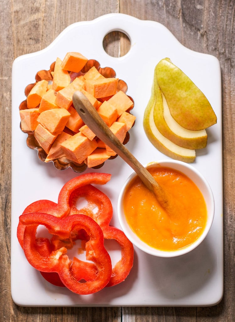 White cutting board with a spread of produce on it with a bowl of smooth baby food.