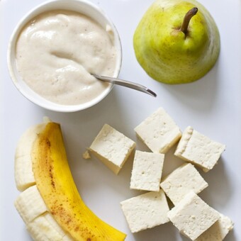 White cutting board with produce spread on it with a small bowl of the baby food puree.