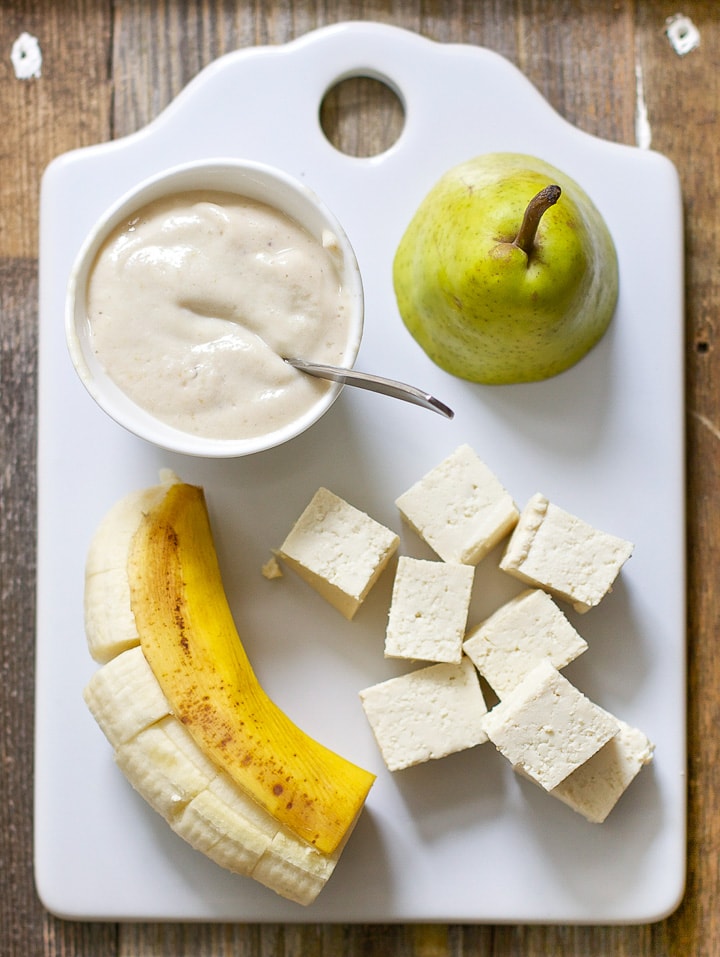 White cutting board with produce spread on it with a small bowl of the baby food puree. 