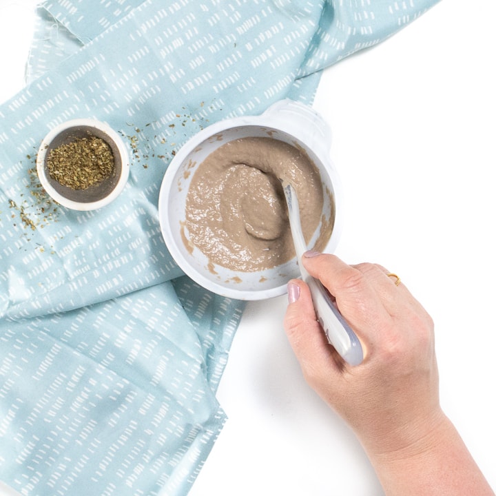 Gray baby bowl with beef pure and a small bowl of dried oregano with hand reaching in to stir the puree.