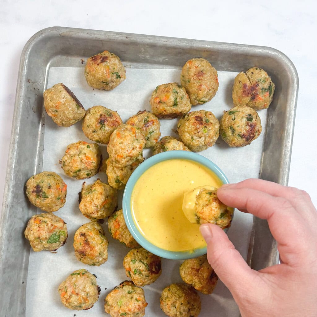 Metal baking tray on my counter, with a hand dipping a meatball into a yellow dip. 