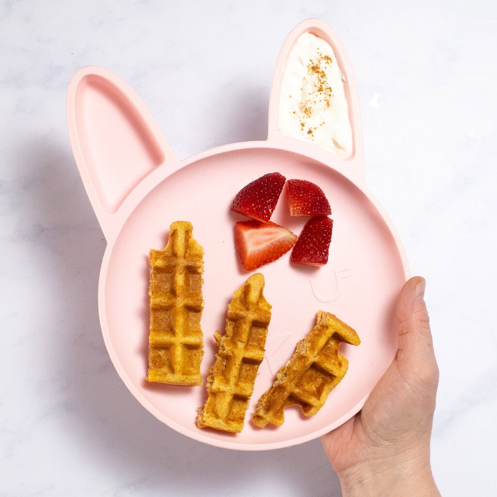 Small pink baby plate in the shape of a bunny with waffles, strawberries and a yogurt and cinnamon dip.