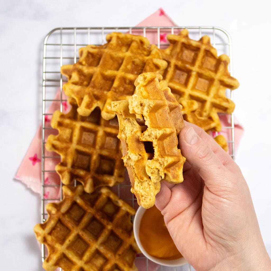 Hand holding a sweet potato waffle over a cooling rack with more waffles and a pink napkin.