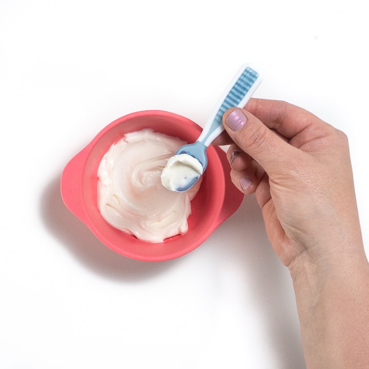 Baby spoon resting on a pink baby bowl with yogurt in it.
