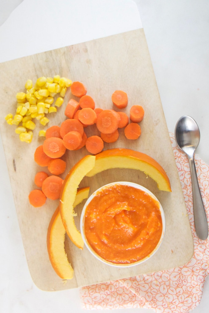 A wooden cutting board with a small white bowl with creamy carrot pumpkin baby food puree with chopped pumpkins, carrots and corn.