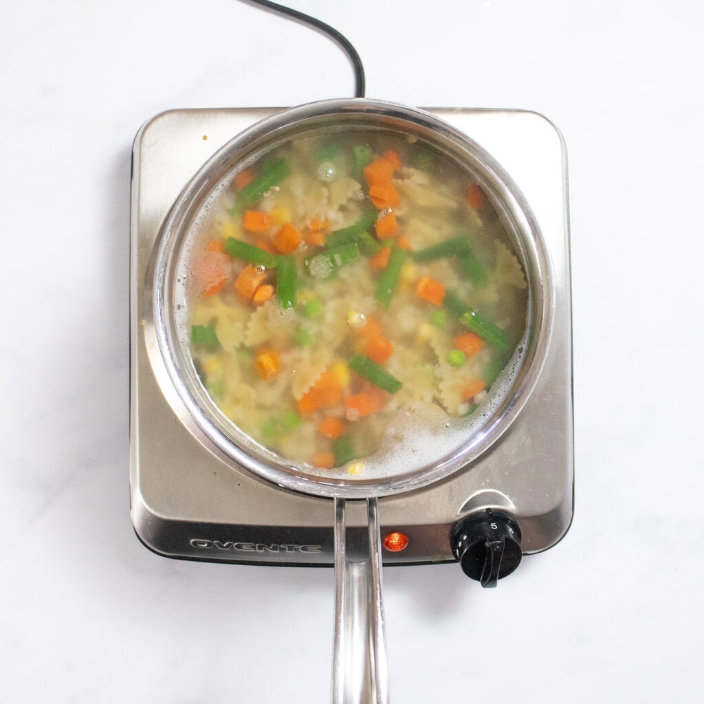 A silver burner with a saucepan with boiling water cooking pasta and veggies.