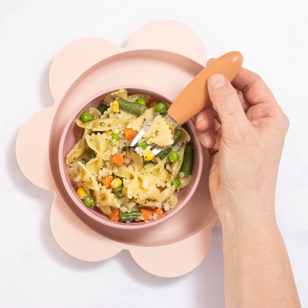 My hand holding a orange baby fork over a bowl which is loaded with cooked pasta and veggies. The bowl is sitting on a pink plate on my marble kitchen counter. 