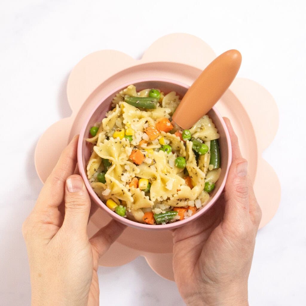 My hands holding a pink kids bowl full of veggie pasta for baby, toddlers and kids over a pink kids plate on my kitchen counter.