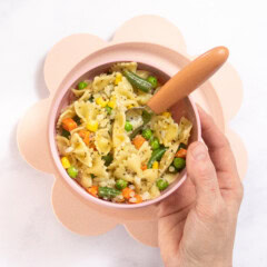 My hand holding a pink baby bowl with a orange baby fork inside, the bowl is full of pasta for baby with a variety of veggies. The bowl is over a pink baby plate that is sitting on my kitchen counter.