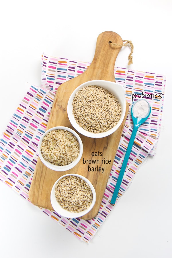 layout of what is in the homemade baby cereal - small white bowls on top of a wood cutting board filled with dry oats, brown rice and barley. On top of a multi colored napkin is a spoon filled with probiotics