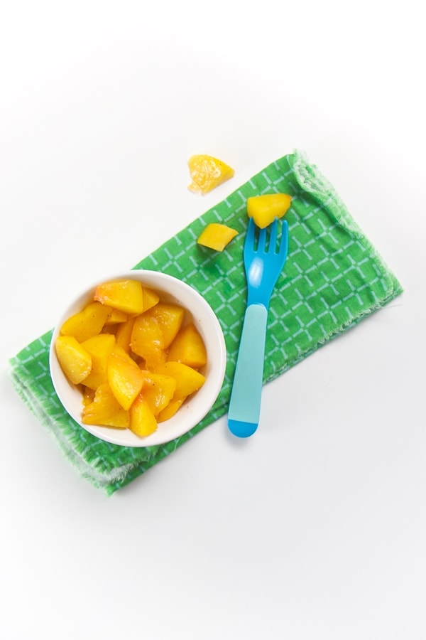 small white bowl on green napkin - finger food of peaches fill the bowl. fork on napkin with a chunk in it.
