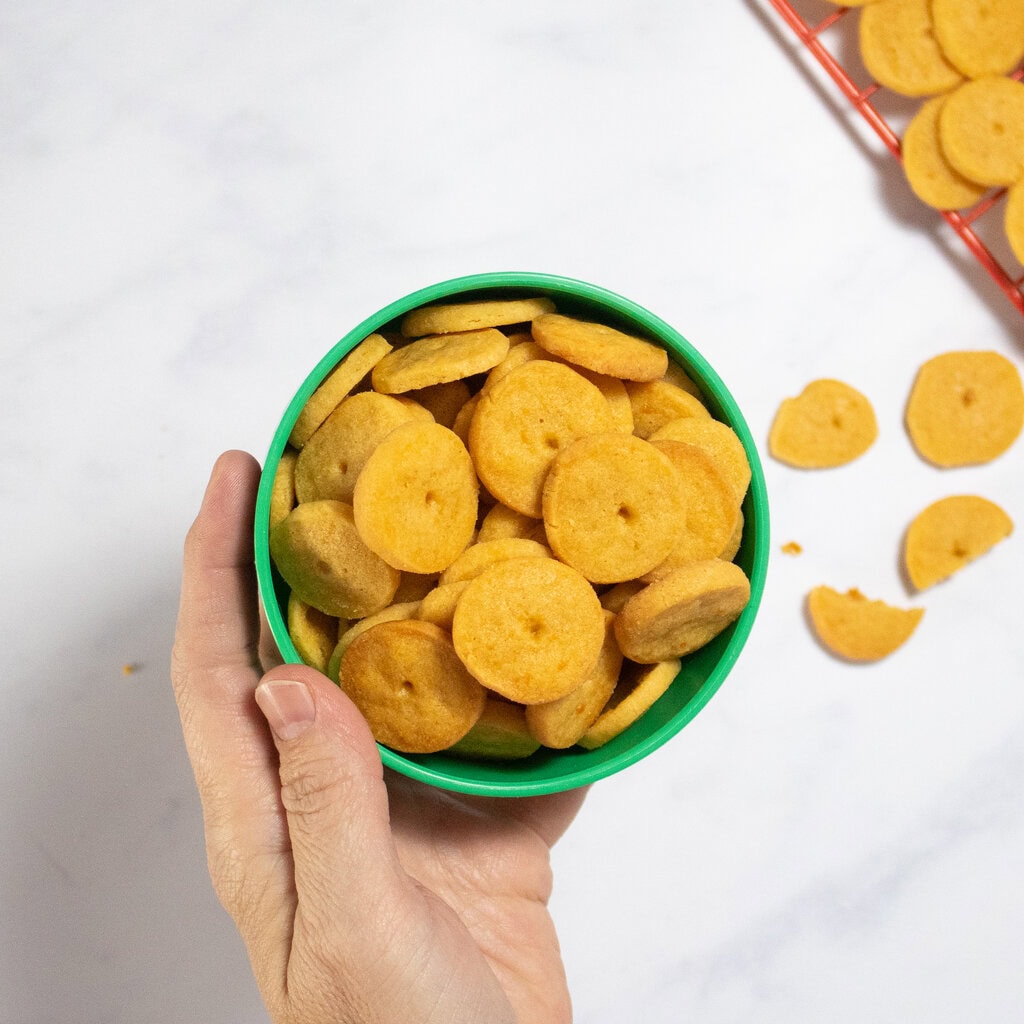 My hand holding a green kids bowl full of golden-brown cheese crackers with a red cooling rack filled with more crackers next to the bowl. 