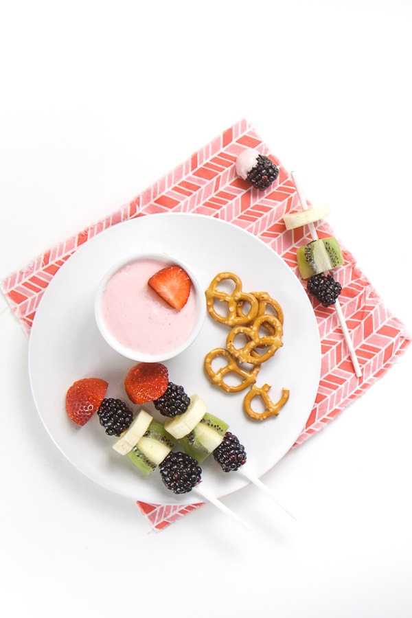 white plate with a small bowl of pink strawberry cream cheese dip and along with 2 fruit kabobs with strawberry, banana, kiwi and blackberry as well as a small pile of pretzels. All on top of a pink napkin.