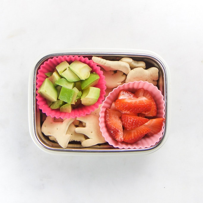 Rectangle kids bento box with healthy snacks for kids - animal crackers and 2 muffin molds - 1 filled with chunks of avocado and the other filled with cut strawberries