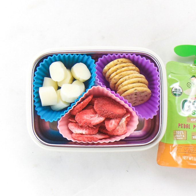 Rectangle kids bento box with healthy snacks for kids - 3 muffin molds filled with cut string cheese, crackers and dried strawberries. On the side is an applesauce pouch