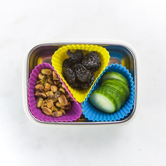Rectangle kids bento box with healthy snacks for kids - 3 muffin molds filled with pistachios, dried fruit and cut cucumber.
