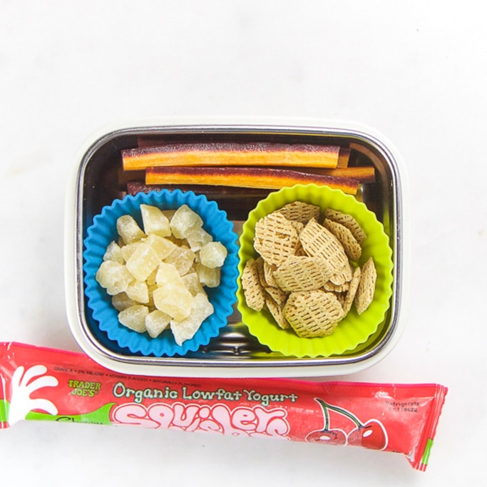 Rectangle kids bento box with healthy snacks for kids - cut purple carrots, dried pineapple and dried cereal and on the side is a frozen yogurt tube.