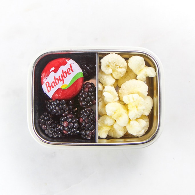 Rectangle kids bento box with healthy snacks for kids - black berries, a babybel cheese circle and half of the container filled with cheese puffs.