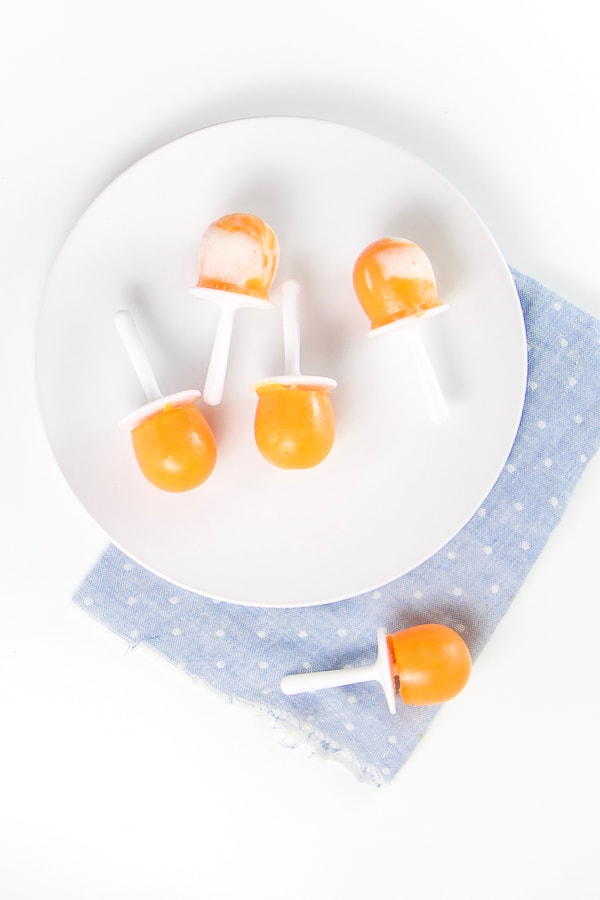 round white plate on top of a blue napkin. On the plate there are 4 orange popsicles with yogurt swirled around in them. there is one orange popsicle on the napkin.