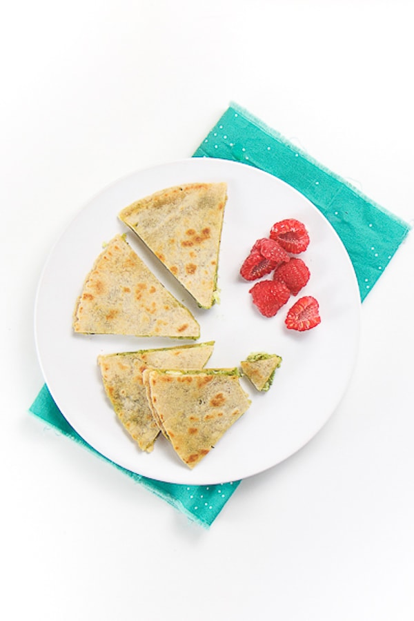 round white plate with wedges of kale pesto chicken quesadilla with a side of chopped raspberries. plate is sitting on a teal napkin on a white surface.