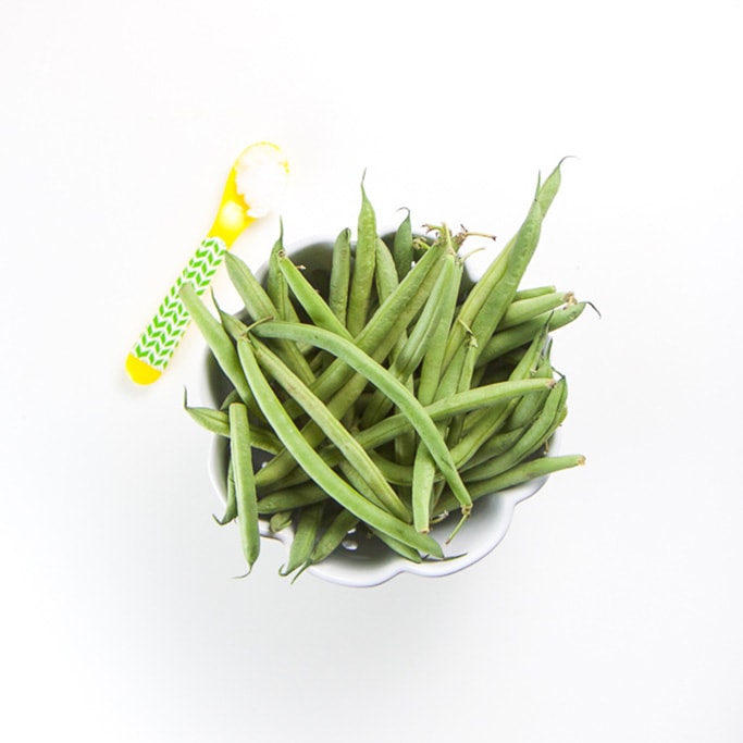small white bowl with scalloped edges sitting on a white background filled with fresh green beans. Yellow and green spoon sitting next to bowl filled with coconut oil