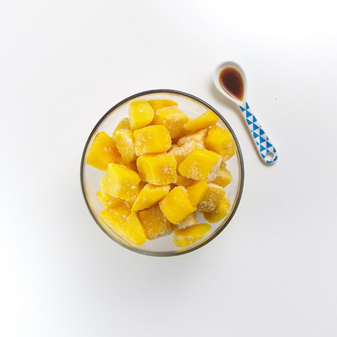 clear glass bowl filled with frozen mango chunks with a small white and blue spoon sitting next to it filled with vanilla bean extract