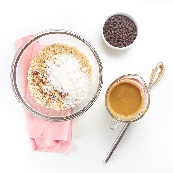 3 bowls and a small whisk sitting on a white background with a pink napkin underneath the biggest bowl. The largest clear glass bowl has the dry ingredients, a small glass measuring cup as the wet ingredients and a small white bowl is filled with mini chocolate chips.