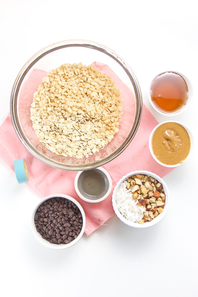 On a white background are 6 bowls scattered around holding the ingredients to make these homemade granola bars - oats, rice crispy cereal, agave nectar, peanut butter, chopped nuts, shredded coconut, coconut oil and mini chocolate chips.
