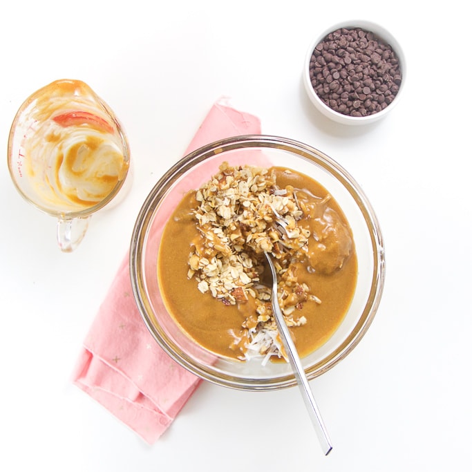 A large clear glass bowl is filled with all of the ingredients for these easy granola bars and is about to get mixed together. there is also a scrapped clean measuring cup and a small bowl of chocolate chips sitting next to the clear bowl.