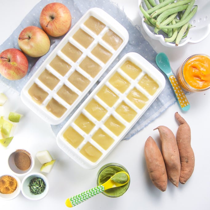 Spread of different produce and jars of organic baby food as well as 2 freezer containers filled with purees sitting on a blue napkin on top of a white backgournd.