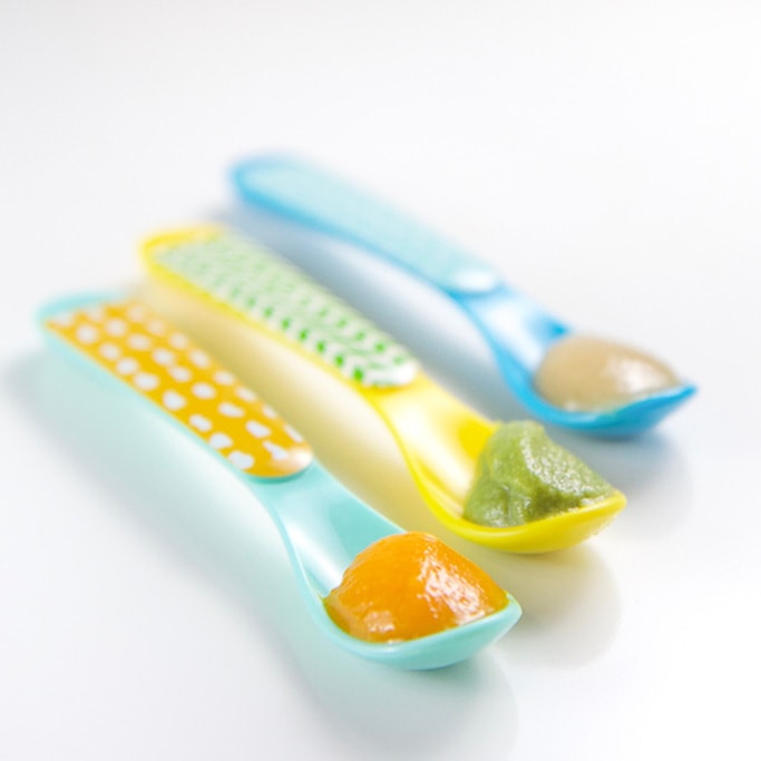 3 colorful spoons sitting on a white background with 3 different organic baby food in them - sweet potato, green bean and apple puree.