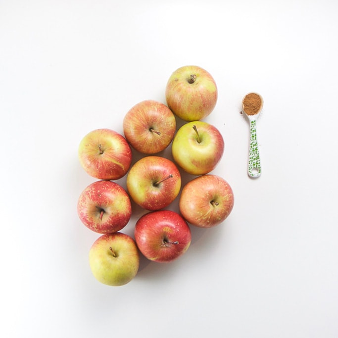 9 apples sitting on a white background in a triangle with a small white and green spoon filled with ground cloves sitting next to the apples.