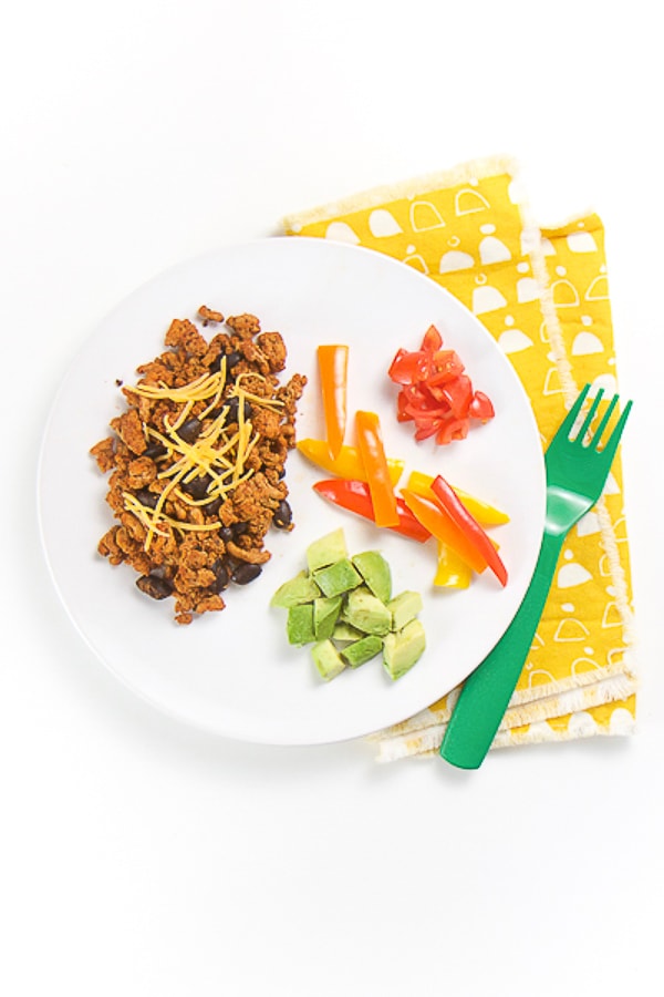round white plate with taco Tuesday baby-led weaning foods on top - ground turkey and beans with a sprinkle of cheese, avocado chunks, pepper strips and tomato chunks