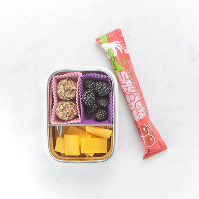 white bento filled with cheese, blackberries and fruit balls with a yogurt squeeze beside it.