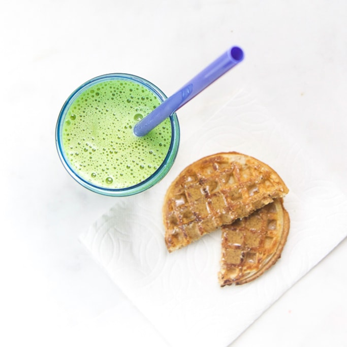 green smoothie in blue toddler cup sitting on white surface with a waffle cut in half on a white napkin.