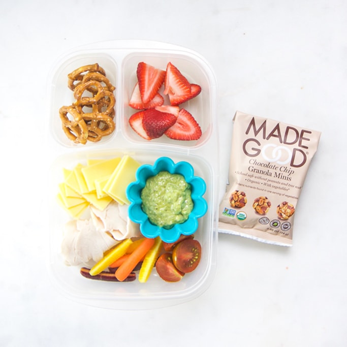 Clear bento no white surface filled with turkey, cheese, carrots and tomatoes with an avocado dipping sauce, chopped strawberries and pretzels with a packed snack.