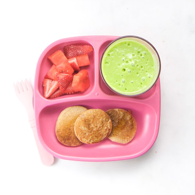 3-section plate on a white surface filled with toddlers breakfast meal - blender pancakes, green smoothie and chopped strawberries.