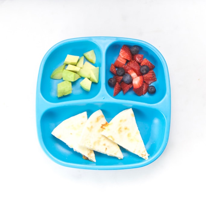 3-section plate on a white surface filled with toddlers lunch meal - cut wedges of bean quesadilla, fruit and avocado.